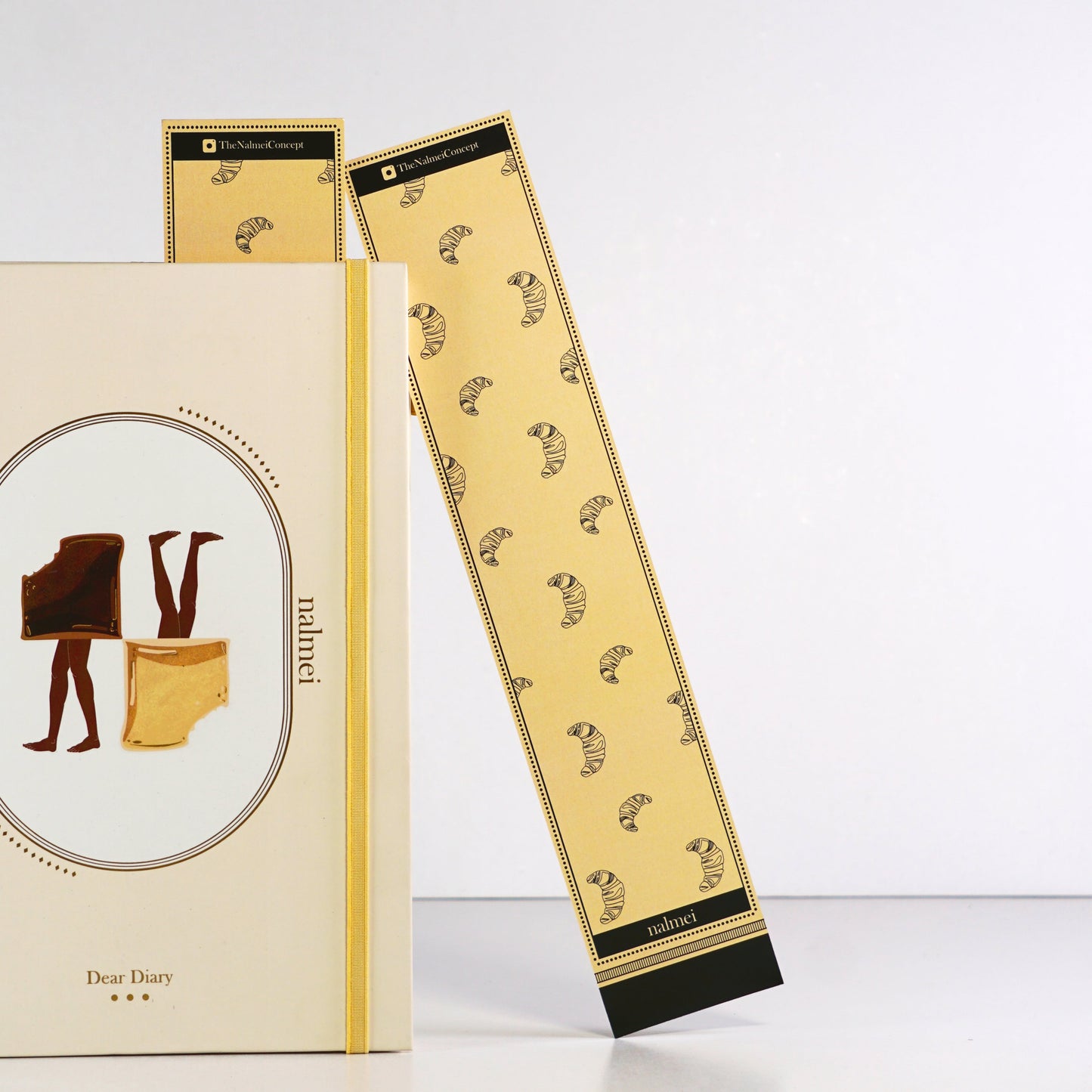 A large beige book mark with black croissant illustrations outlined | a bookmark leaning on a quotes journals | Nalmei